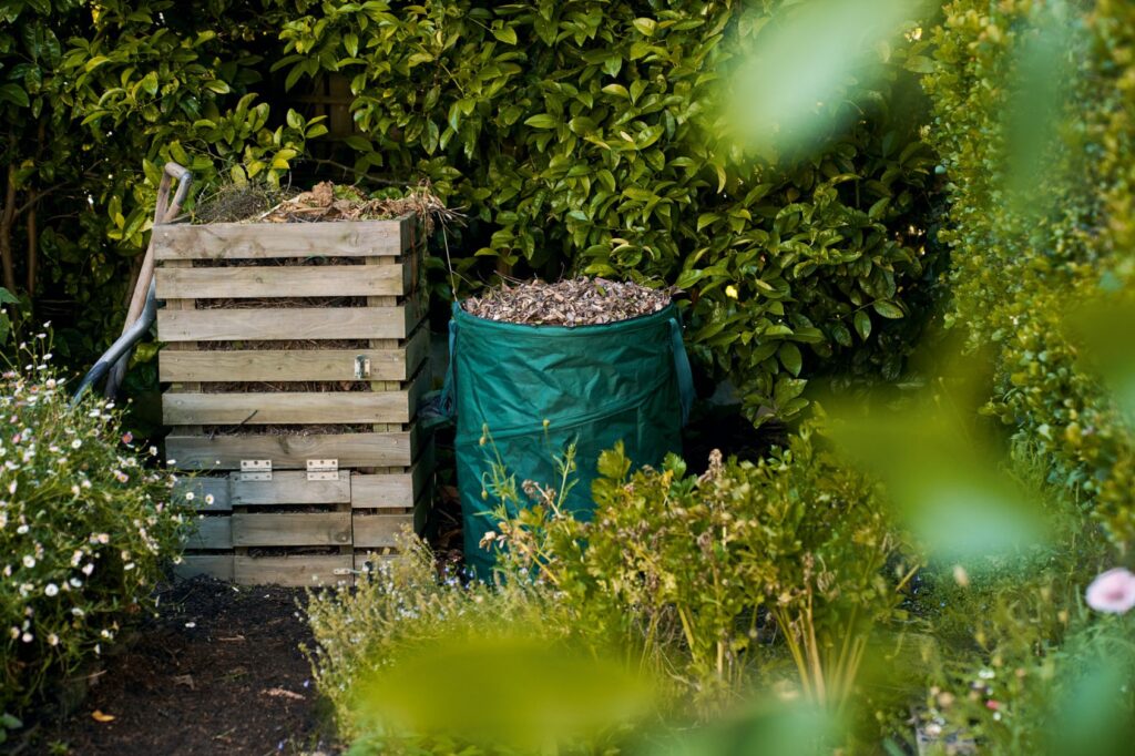 Tuinafvalzak naast een houten compostbak vóór een groene haag